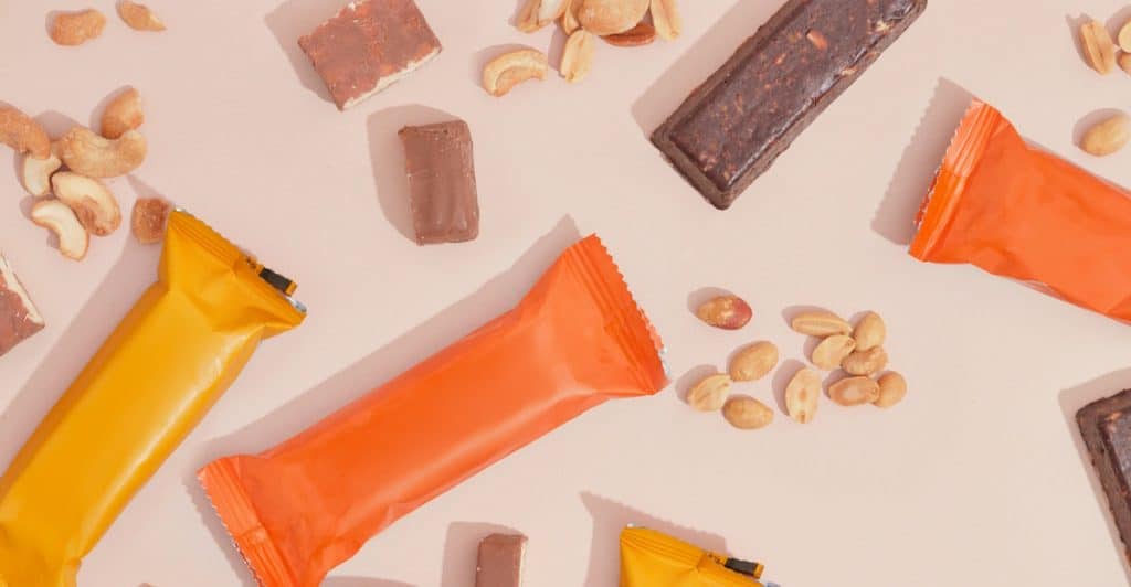An overhead shot of various protein bars, nuts, and chocolate pieces scattered on a light pink surface.