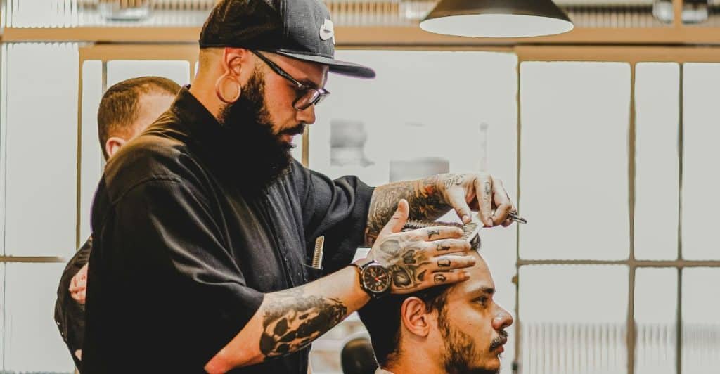 A barber with tattoos and a black cap is cutting a man's hair in a barbershop.