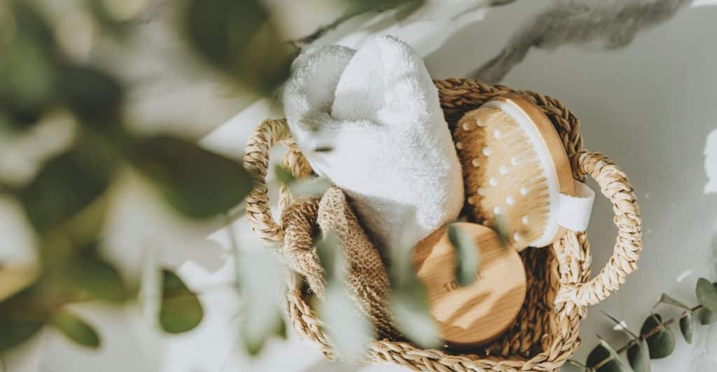 A basket containing shower essentials like slippers and a brush, with green leaves in the foreground.