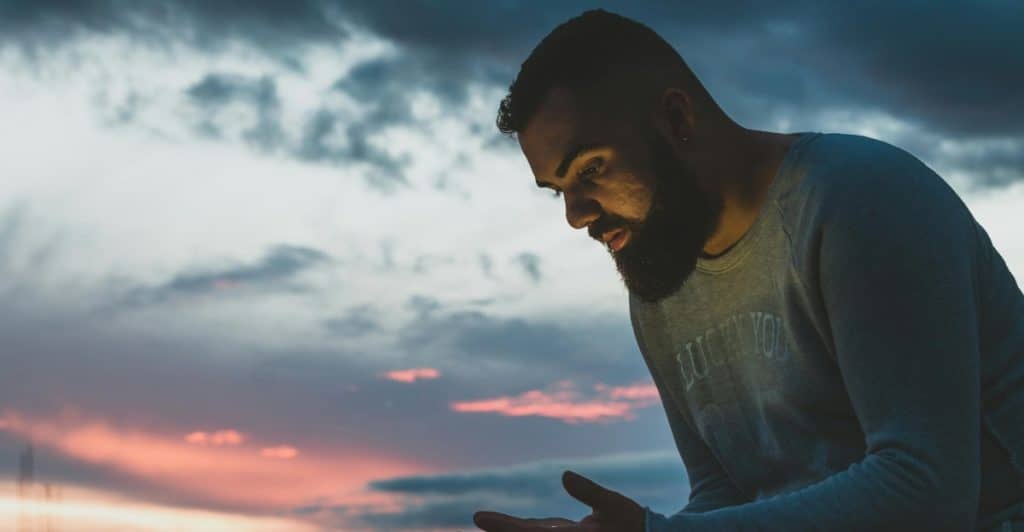 A bearded man sitting outdoors at sunset looking at his hands.