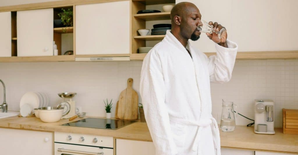 A Black man in a white bathrobe is standing in a kitchen and drinking a glass of water.