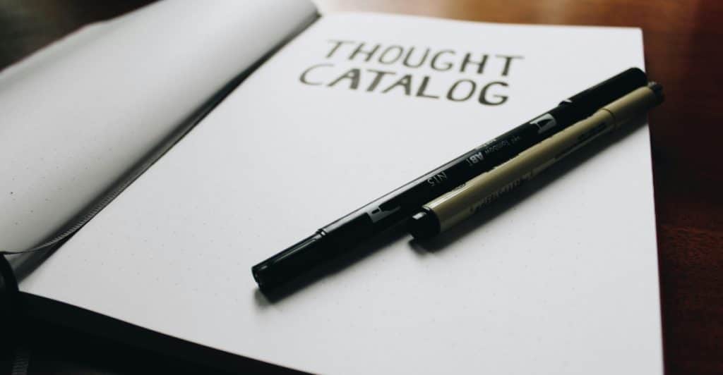 Close-up of an open notebook with the words "THOUGHT CATALOG" written on the cover, with two pens lying on the pages.