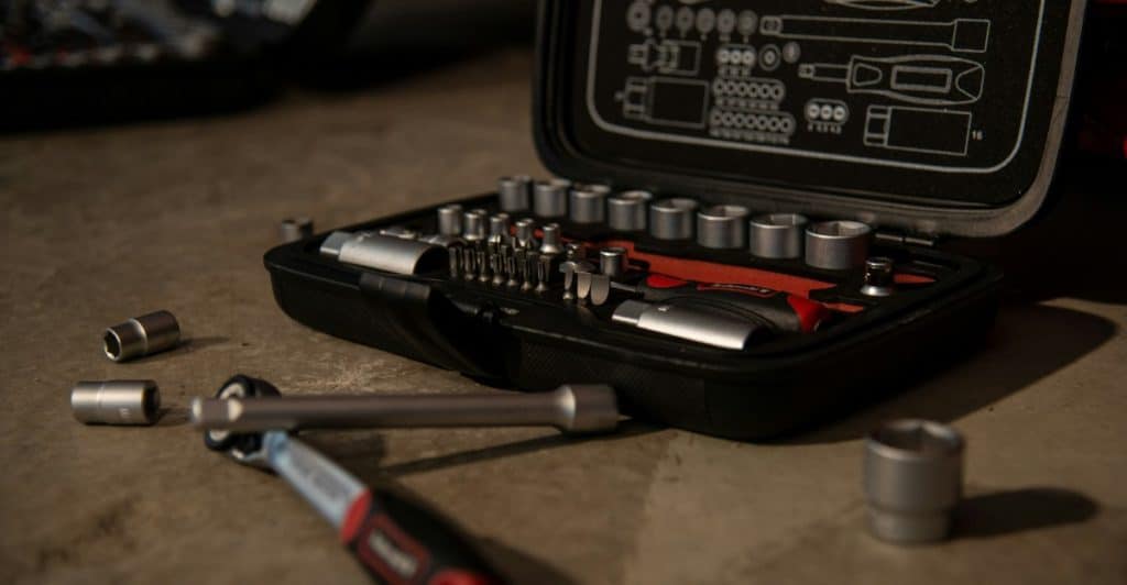 A black toolbox kit is open on a concrete floor, revealing various metal sockets, wrenches, and screwdriver bits.