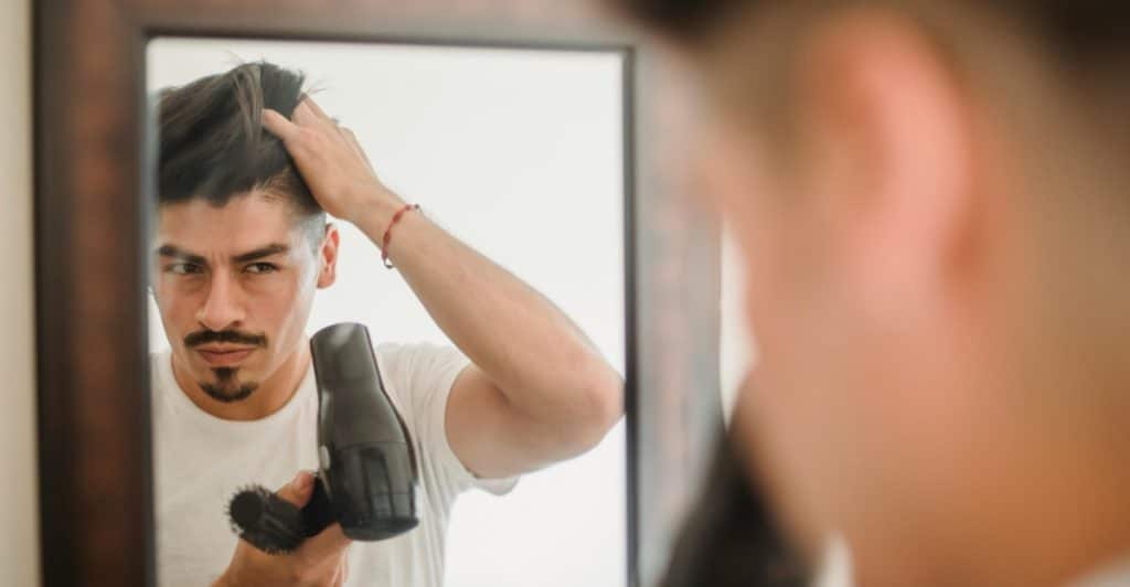 A man is looking into a mirror and using a blow dryer on his hair while holding a brush.