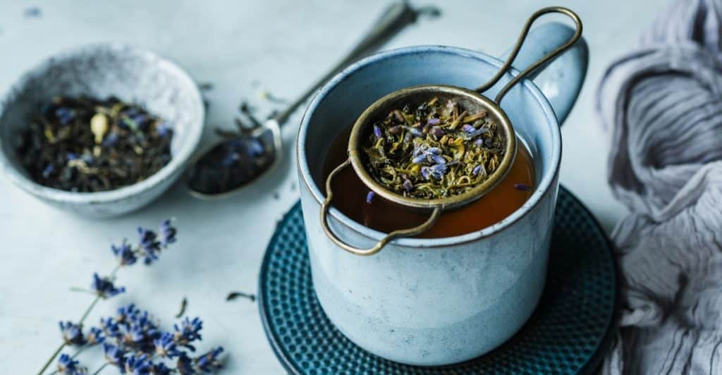 Blueberry leaf tea being strained into a blue mug with lavender flowers nearby.