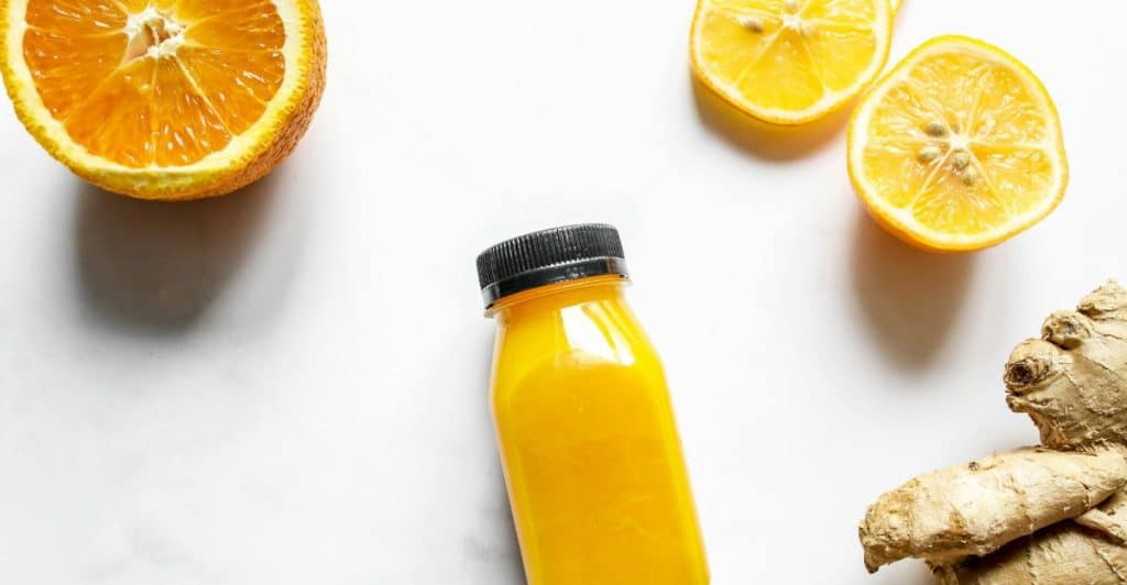 A bottle of ginger lemonade on a white surface, surrounded by orange slices and ginger root.