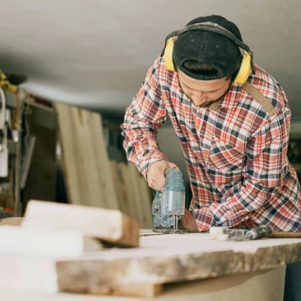 man wearing a plaid shirt and yellow ear protection uses a jigsaw to cut a piece of wood in a workshop.