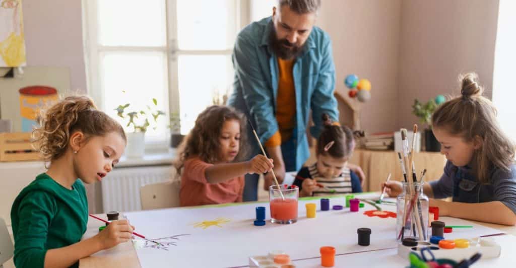 Several young children painting on a large sheet of paper at a table with the help of an adult.