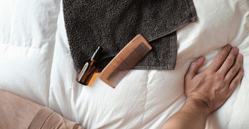 A hand reaching towards a comb and small bottle on a white surface next to a folded towel.