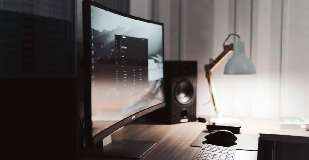 A curved monitor, keyboard, mouse, and lamp on a wooden desk.