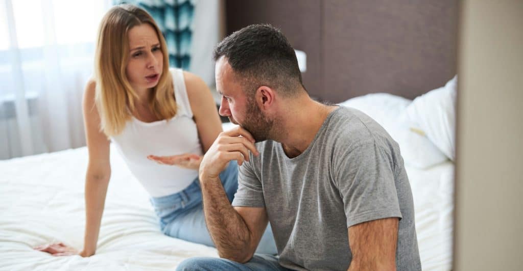A couple in a bedroom, with one person sitting on the bed gesturing and the other listening with a concerned expression.