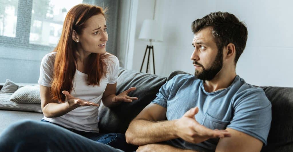 A couple sits on a couch, gesturing and looking upset while in discussion.