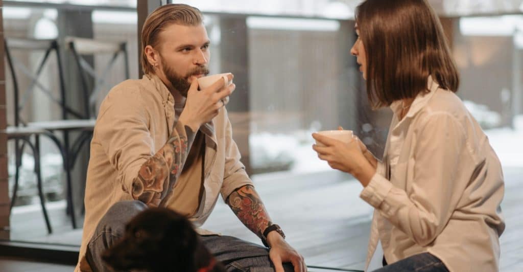  A man and a woman sitting on the floor, drinking coffee, with a small dog nearby.