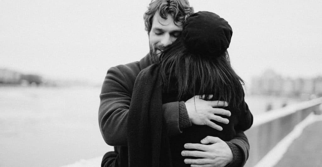 A black and white image of a couple embracing outdoors near a body of water.