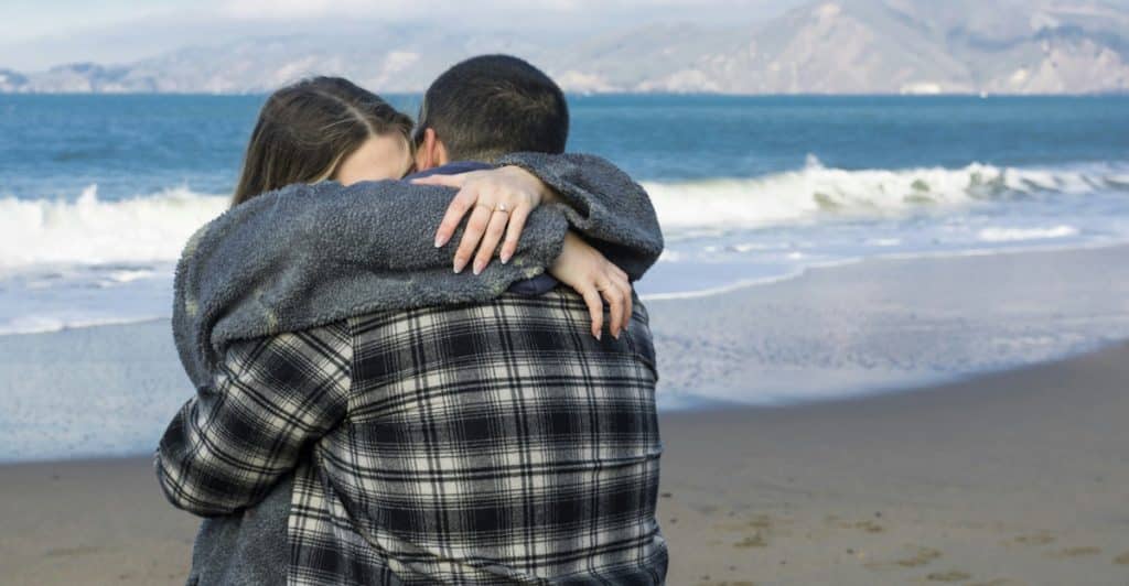 A couple embracing on a beach, with an ocean and hilly landscape in the background.