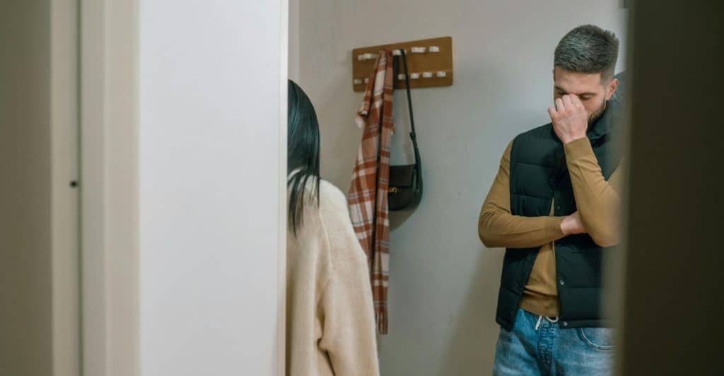 A man covering his face with his hand while standing facing away from a woman in a hallway, suggesting conflict or disagreement.