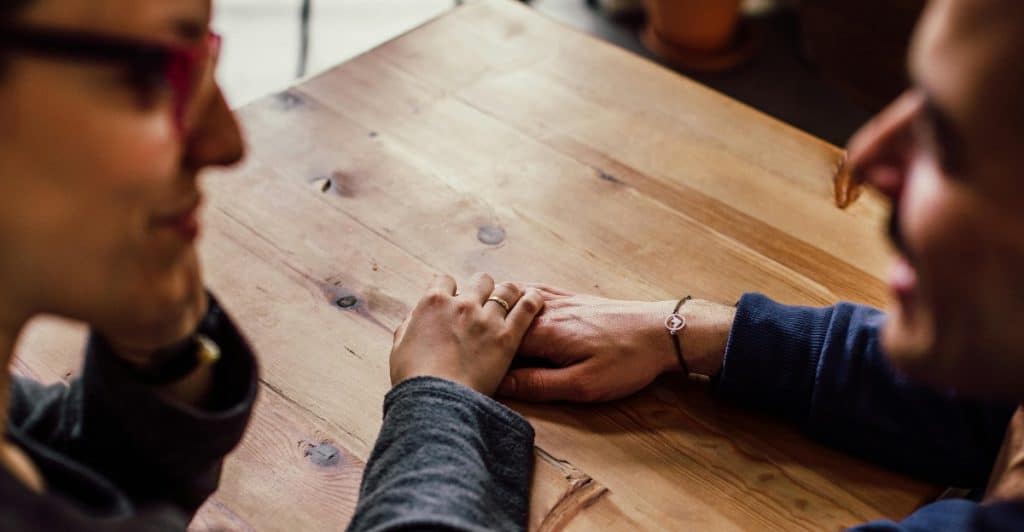 Two people holding hands across a wooden table, seen from the shoulders up, suggesting close communication.