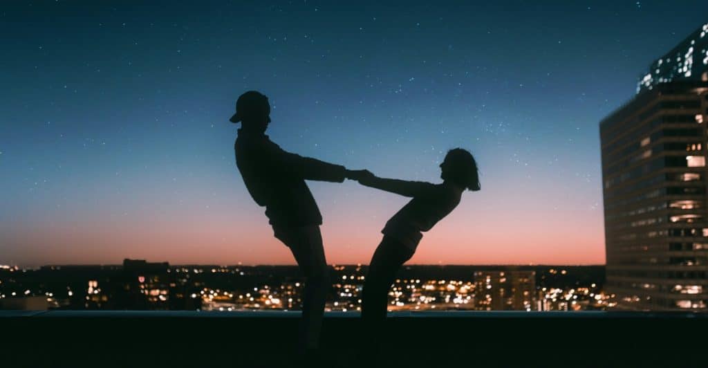 Two silhouetted figures holding hands and leaning away from each other on a rooftop at sunset, with city lights in the background.