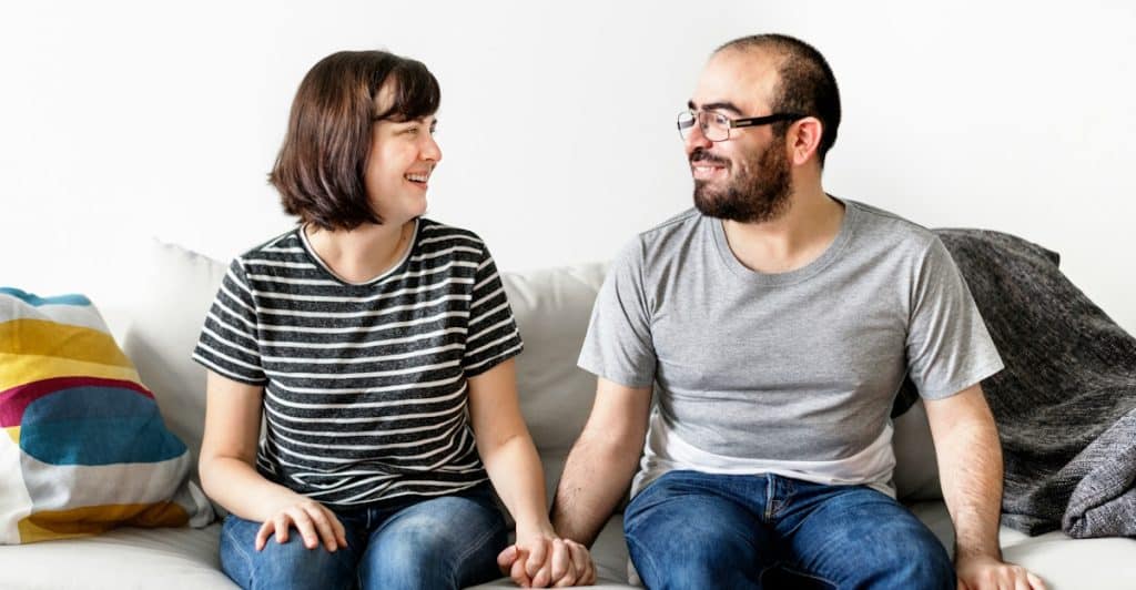 A couple sitting together on a couch, holding hands and smiling at each other.