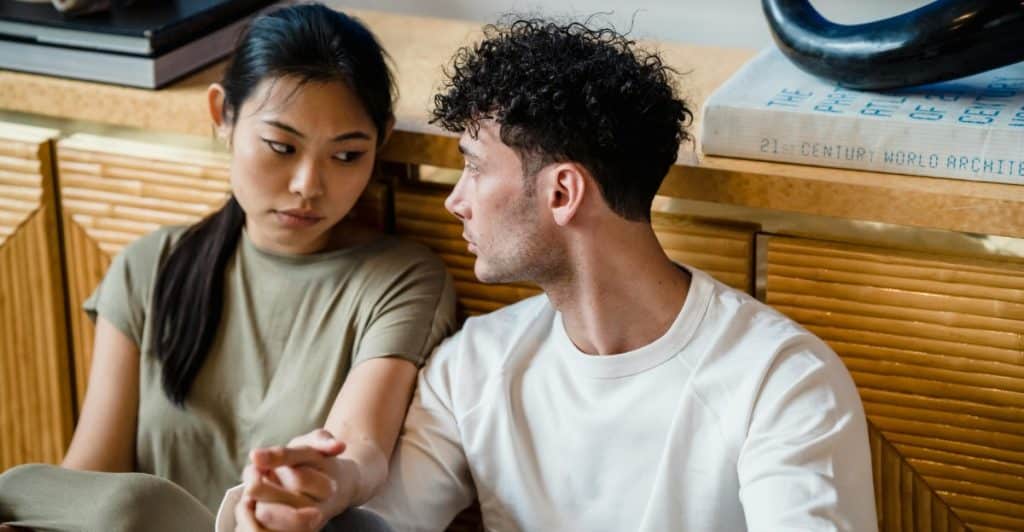 A man and a woman sitting close together, holding hands, with serious expressions.