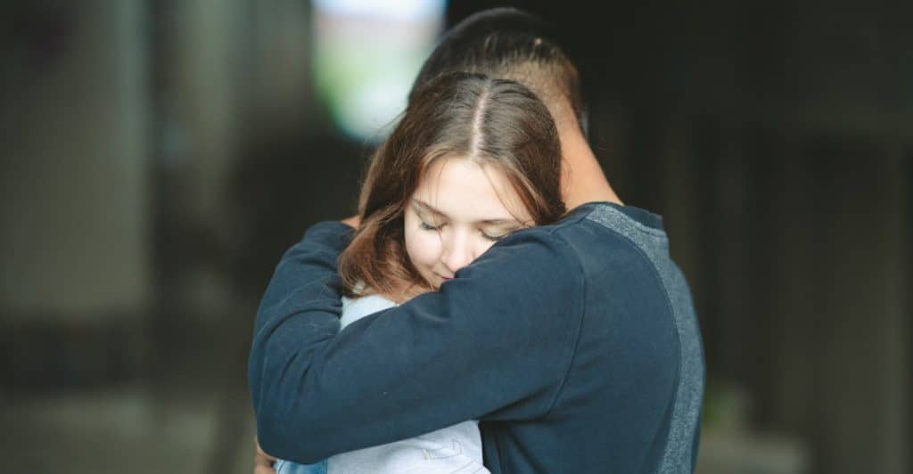 A woman with long brown hair is embracing a man, burying her face into his shoulder.