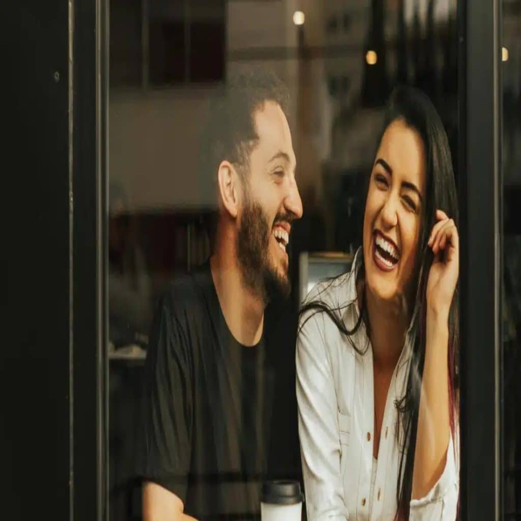 Laughing couple sitting by a glass window in a cafe with a coffee cup on the table.