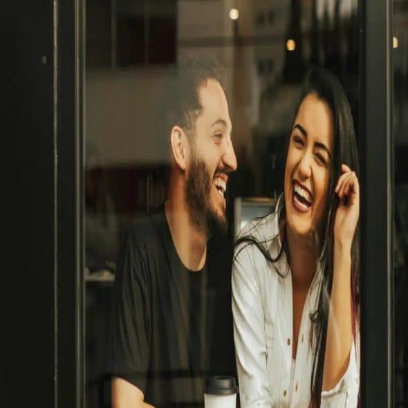 Laughing couple sitting by a glass window in a cafe with a coffee cup on the table.