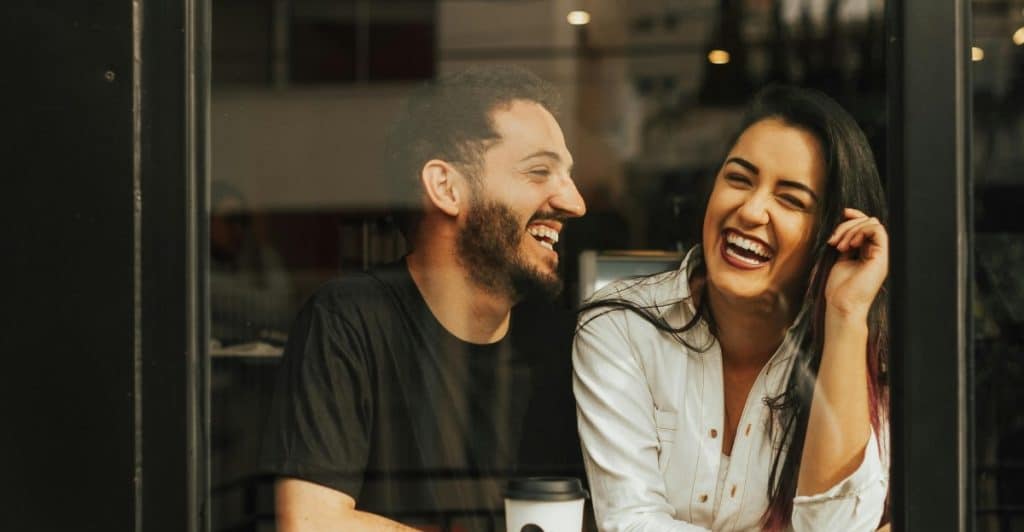 Laughing couple sitting by a glass window in a cafe with a coffee cup on the table.