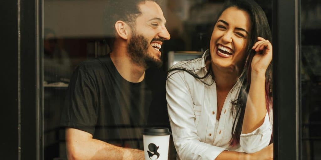 Laughing couple sitting by a glass window in a cafe with a coffee cup on the table.