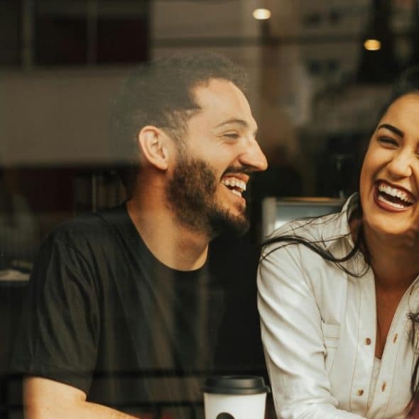 Laughing couple sitting by a glass window in a cafe with a coffee cup on the table.