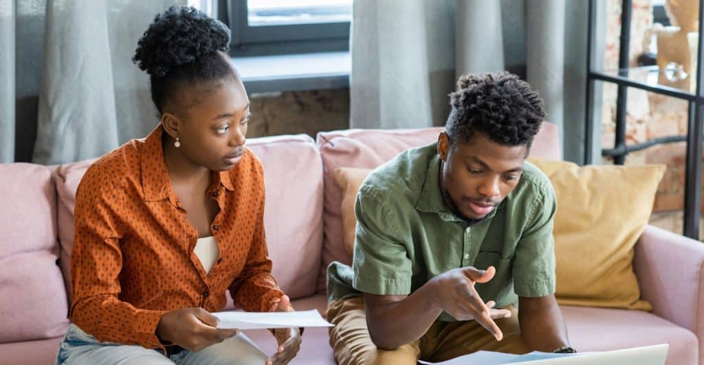 A couple sits on a pink couch, reviewing papers and a laptop together.