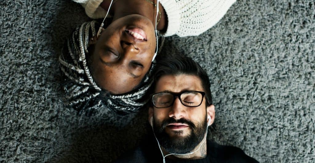Two people lying on a carpet, head-to-head, with eyes closed and sharing earphones, enjoying music together.