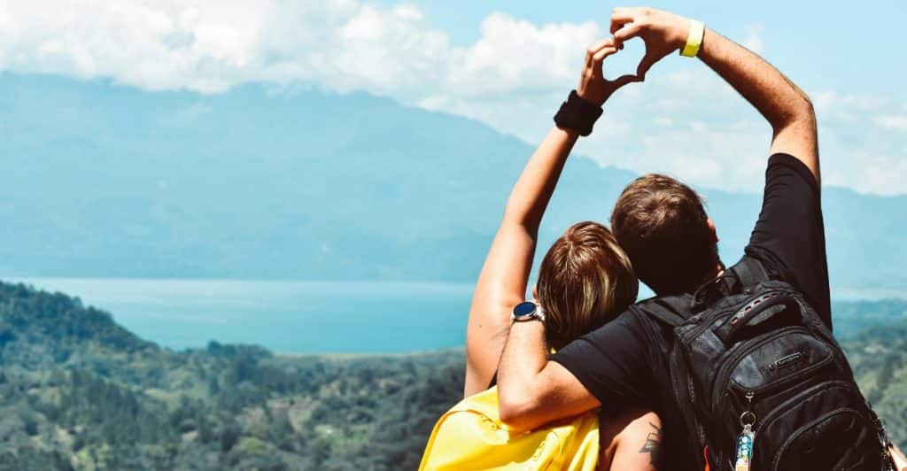 A couple with backpacks standing on a hill making a heart shape with their hands against a scenic background.