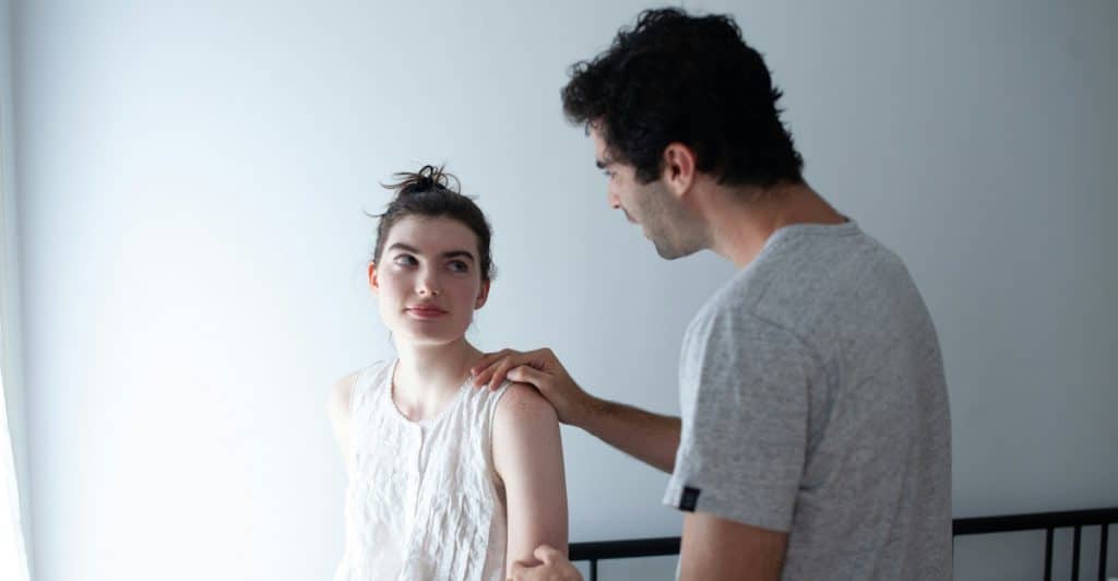 A man in a grey shirt with his right hand on the shoulder of a woman in a white dress in a bedroom setting.