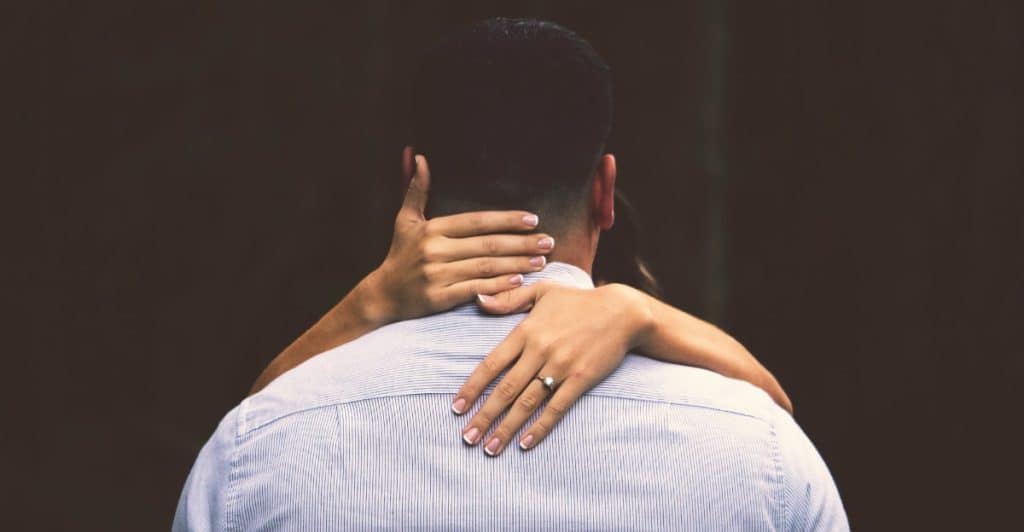 A close-up from behind a man, showing a woman's arms wrapped around his neck with her hands clasped, highlighting connection and respect for boundaries.