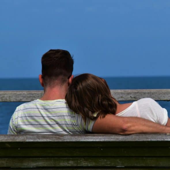 A couple sits closely together on a wooden bench, facing away from the viewer, with a blue sea and sky in the background.