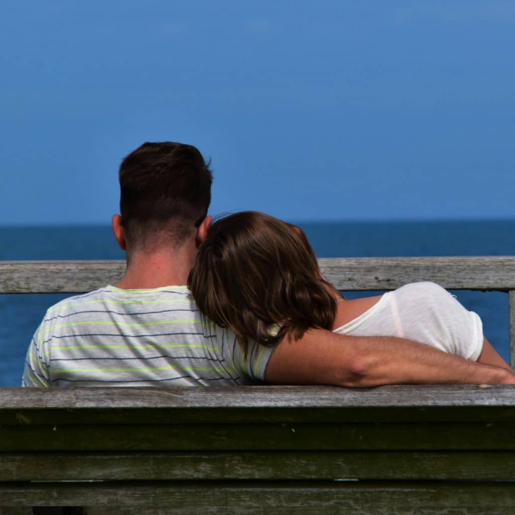 A couple sits closely together on a wooden bench, facing away from the viewer, with a blue sea and sky in the background.