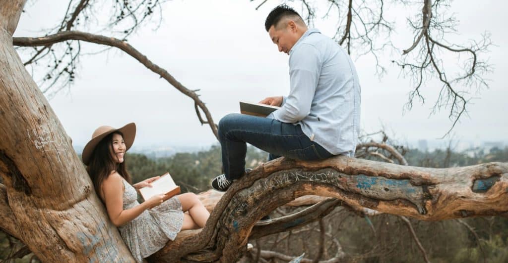 A couple sitting on the large branch of a tree outdoors, reading books and looking at each other, with a blurred landscape in the background.