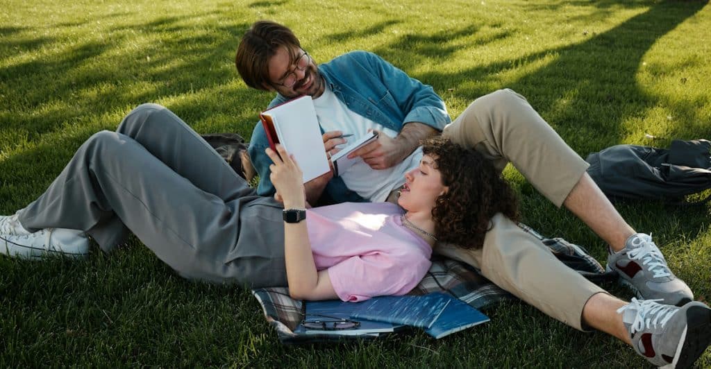 A couple is lying on a blanket in a grassy area, looking at a book and papers.