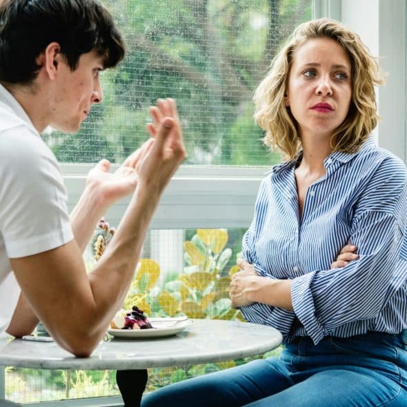 A man and a woman seated at a table in what appears to be a cafe or sunroom, engaged in a serious conversation.