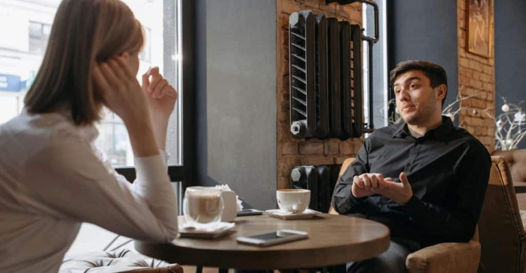A man and a woman are seated at a table in a cafe, engaged in a serious conversation