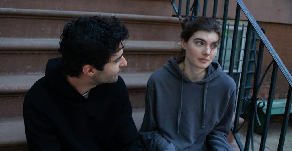 A couple is seated on outdoor steps, engaged in a conversation with serious expressions.