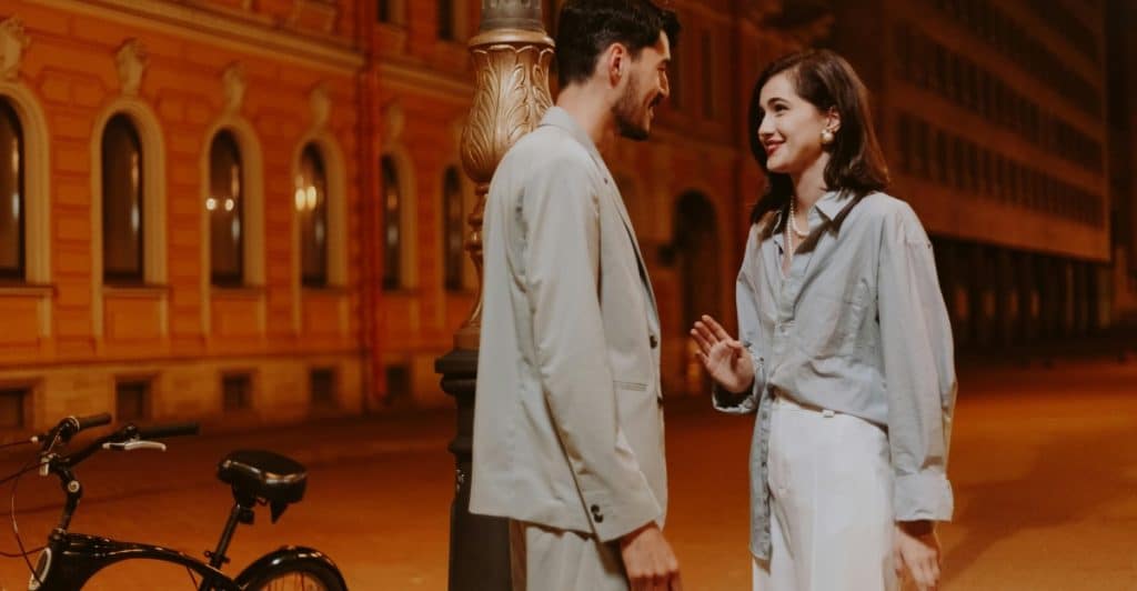  A man and a woman are standing on a city street at night, engaged in conversation.