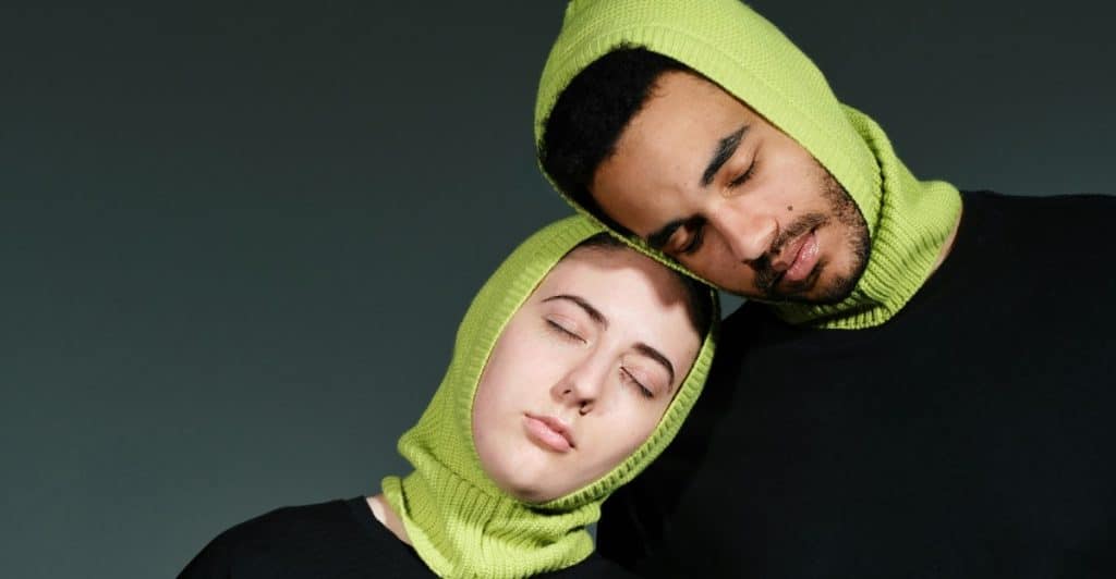 Two people wearing matching green balaclavas, leaning their heads together with their eyes closed, suggesting closeness and growing together.