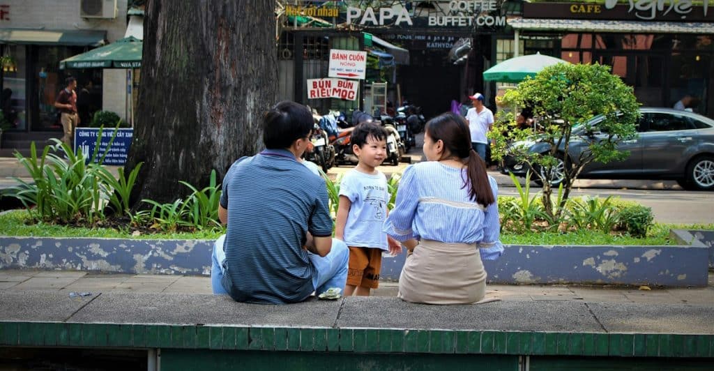 An Asian couple with their toddler sitting outdoors overlooking a street and shops.