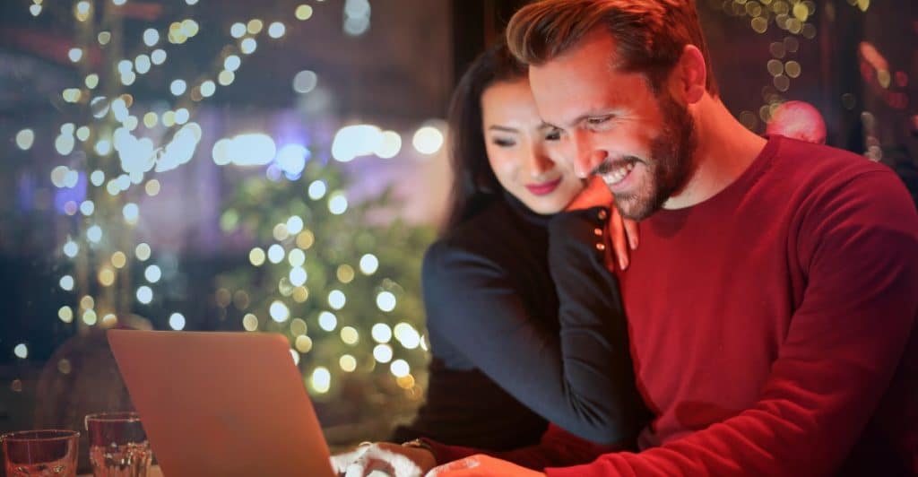 A couple smiles while looking at and typing on a laptop together at a table, with blurred festive lights in the background.