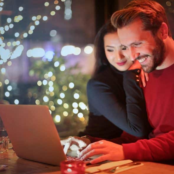 A couple smiles while looking at and typing on a laptop together at a table, with blurred festive lights in the background.