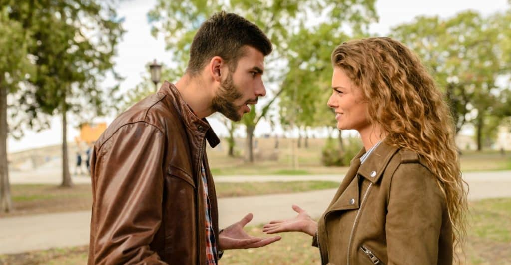 A couple in brown jackets standing facing each other outdoors, gesturing and appearing to have a disagreement.