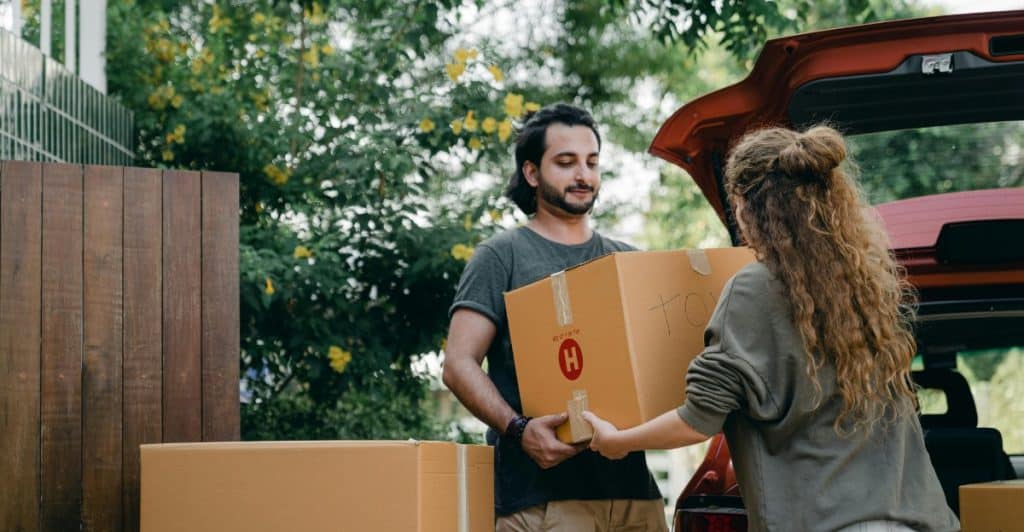 A couple carrying a cardboard box towards the open trunk of a car outdoors, surrounded by greenery.