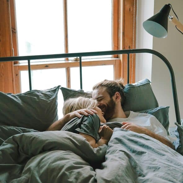 A couple lying in bed, facing each other and smiling, with natural light coming through a window.